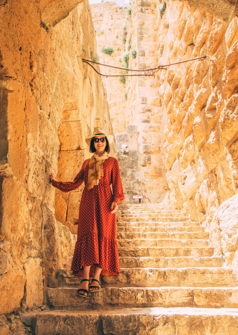 A woman poses on stone steps in Jordan. She's wearing a long red dress, with long sleeves and white polka dots, with sandals, a scarf, and sun hat.