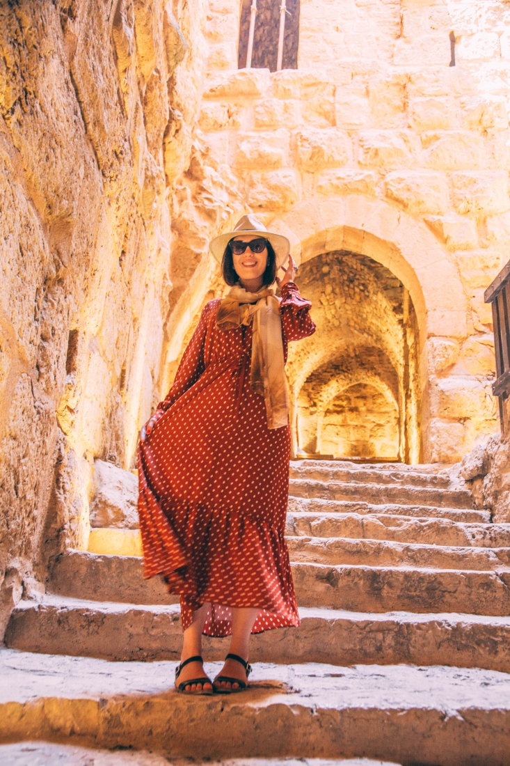 A woman poses on stone steps, her long red maxi dress flowing in the breeze. The dress has long sleeves and small white polka dots. She's wearing a scarf, hat, and sunglasses and smiling at the camera.