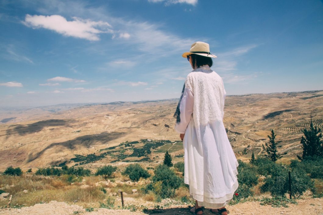 A woman in a long white duster standing, looking out across a mountainous landscape in Jordan