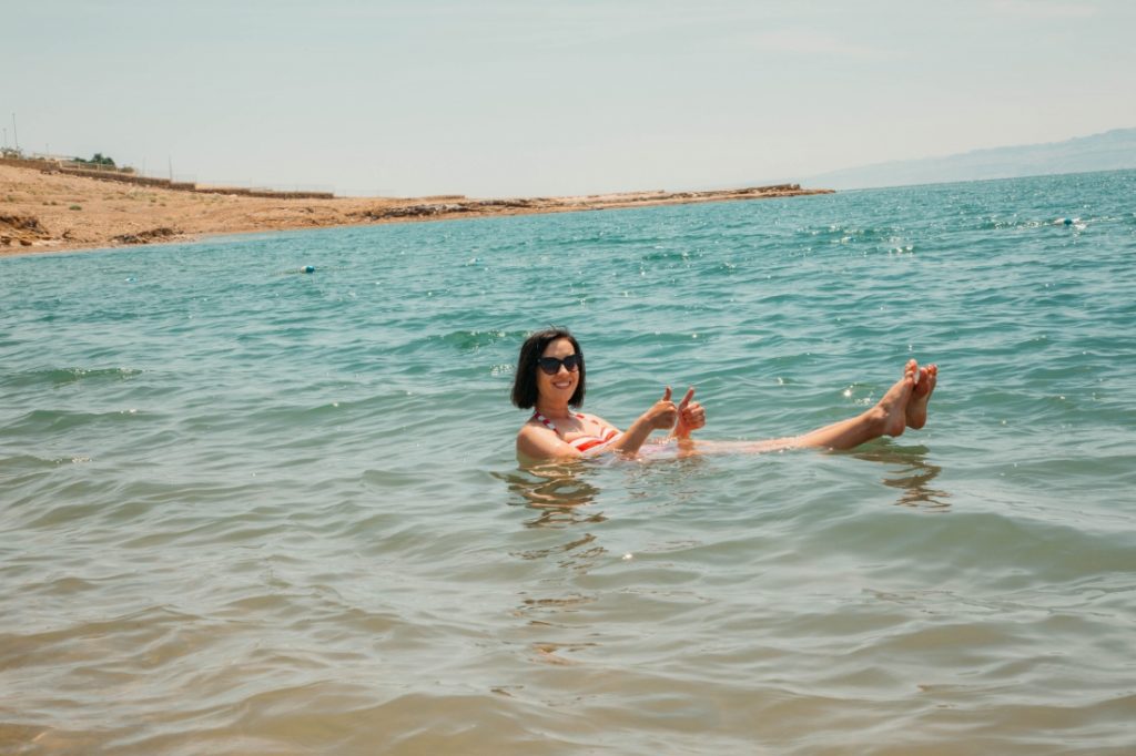 A woman with short brown hair and sunglasses floats in blue murky water. Her head. shoulders, and feet float about the surface of the water.