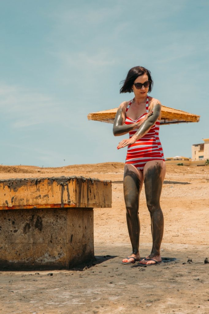 A young women stands next to a large box filled with mud. She's wearing a white and red striped swimsuit and has Dead Sea mud spread all over her arms and legs.