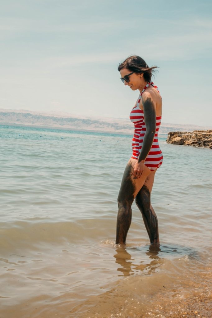 A woman wades into ankle-deep water on the shoreline of the Dead Sea. The water is a murky brown. She's wearing a red and white stripped one-piece swimsuit and has Dead Sea mud spread on her arms and legs.