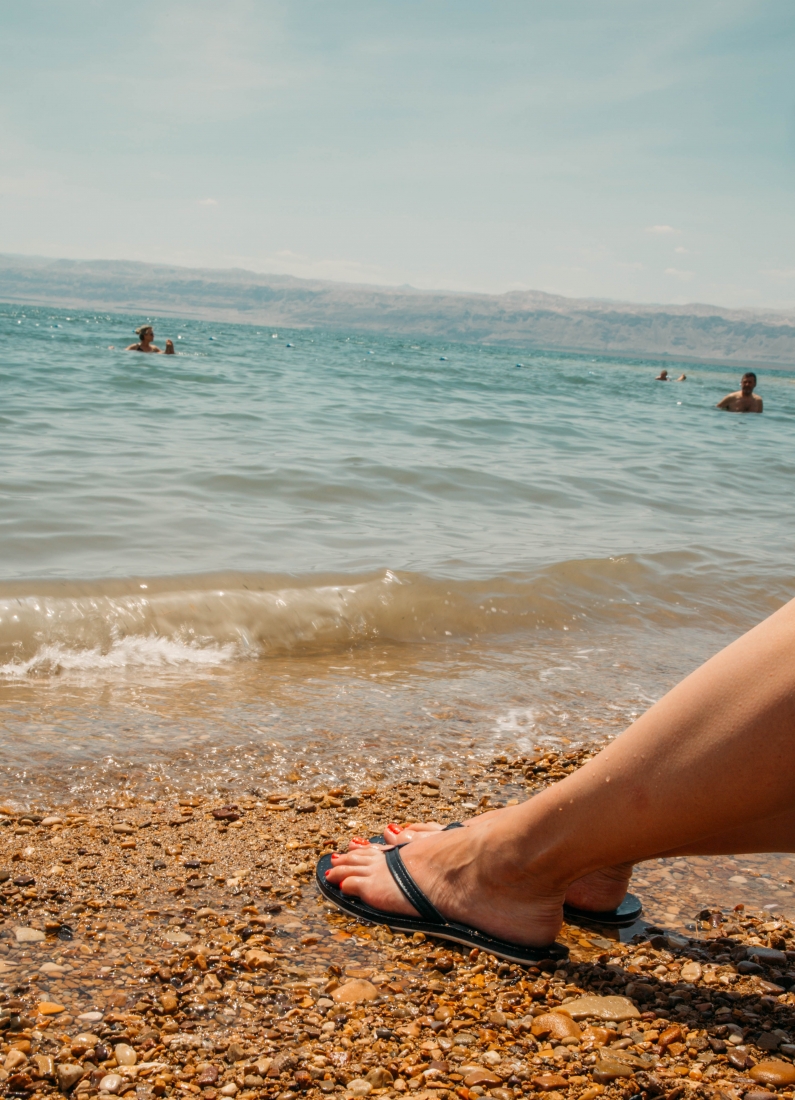 A close up image of a woman's feet on a pebbled beach. She's wearing black flip flops.