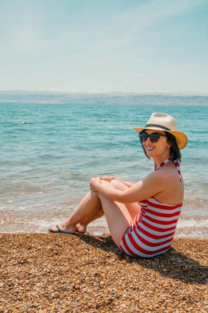 A young woman sits on the pebbled shoreline of the Dead Sea in Jordan, her feet dipped in the crystal blue water. She's wearing a red and white stripped swimsuit, sunglasses, and a hat, and is smiling as she looks over her shoulder.