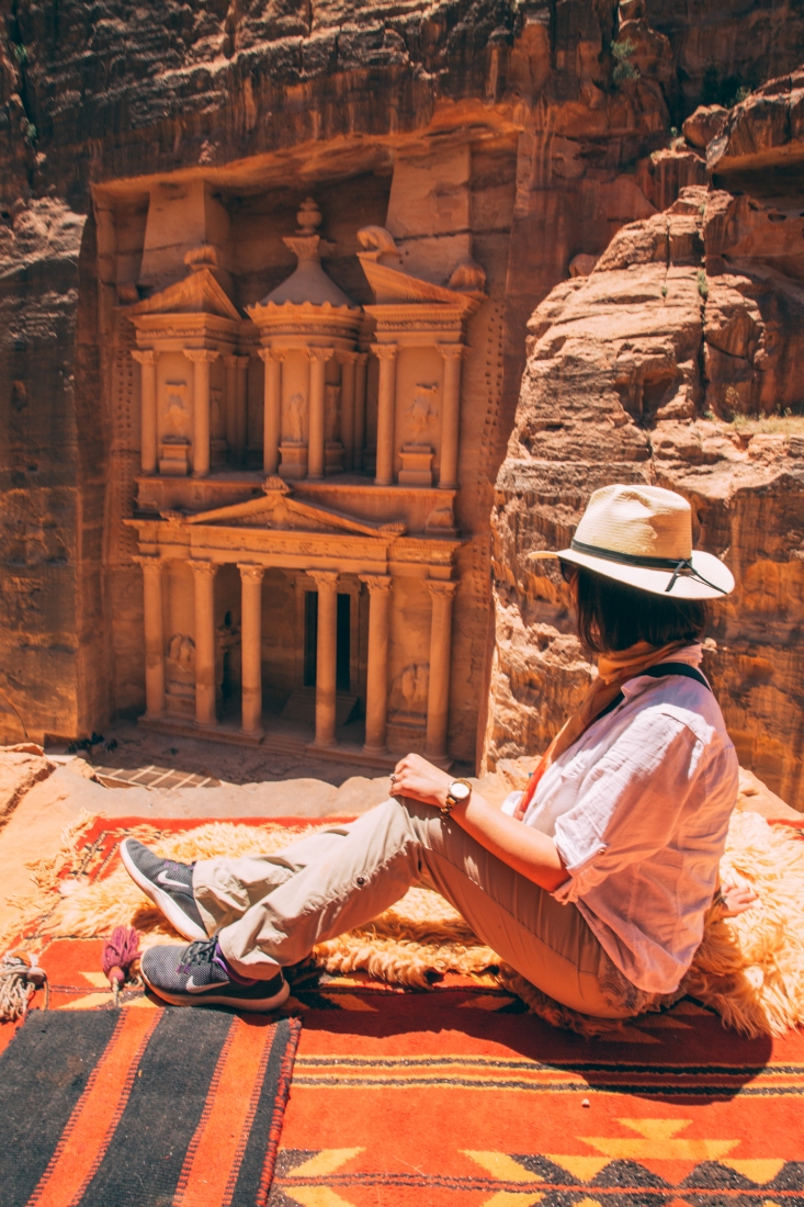 Lindsey of Have Clothes Will Travel wearing Nike shoes for wide feet, Eddie Bauer hiking pants, a straw fedora and a white button down shirt looking over at the Lost City of Petra in Jordan