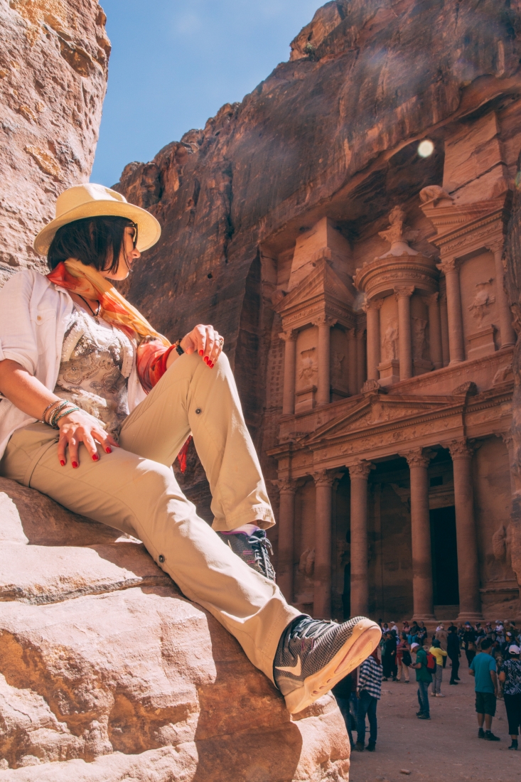 A woman sitting on a rock, wearing hiking pants and sneakers, looking over her shoulder at the Treasury in Petra