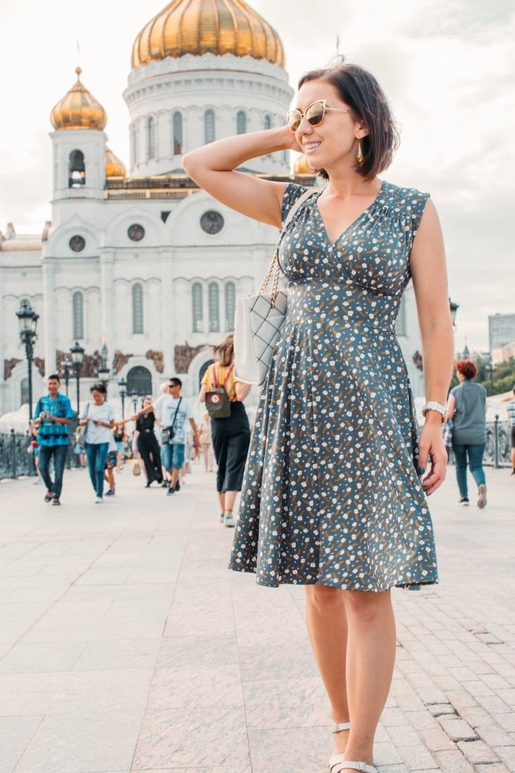 A woman poses in front of a large Russian building as people walk around in a large courtyard. She runs her hands through her short brown hair. She's wearing a blue dress with an all over clover flower pattern.