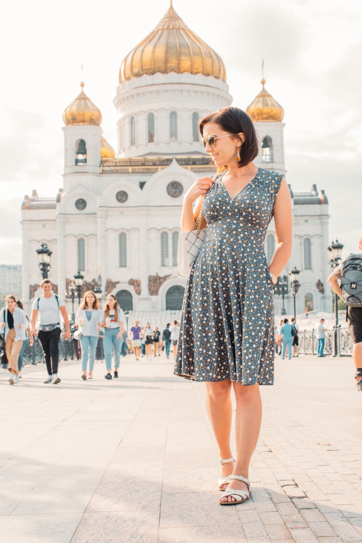 A woman poses in front of a giant Russian building in a courtyard full of people. She's wearing a knee-length dress with an all-over clover flower pattern, wearing sunglasses and sandals.