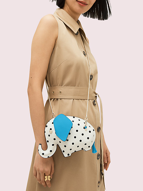 A model wears a small crossbody bag shaped like a small elephant. The elephant purse is white with black polka dots and bright blue ears.