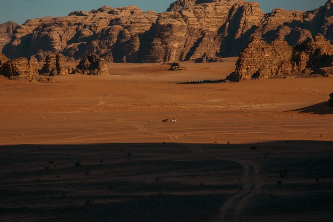 An expansive landscape view of the red sands and rocky mountains of the Wadi Rum desert. In the center of the image, far off in the distance, is a couple riding horses through the desert.