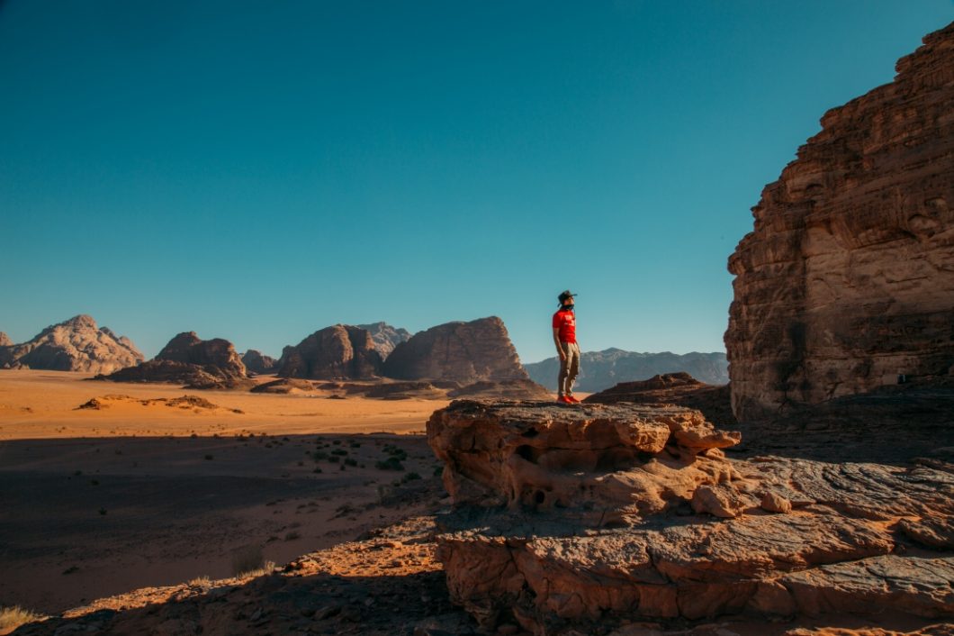 A man stands atop a low rock formation in the middle of the Wadi Rum desert. He's surrounded by vast sandy valleys and rocky mountains under clear blue skies.