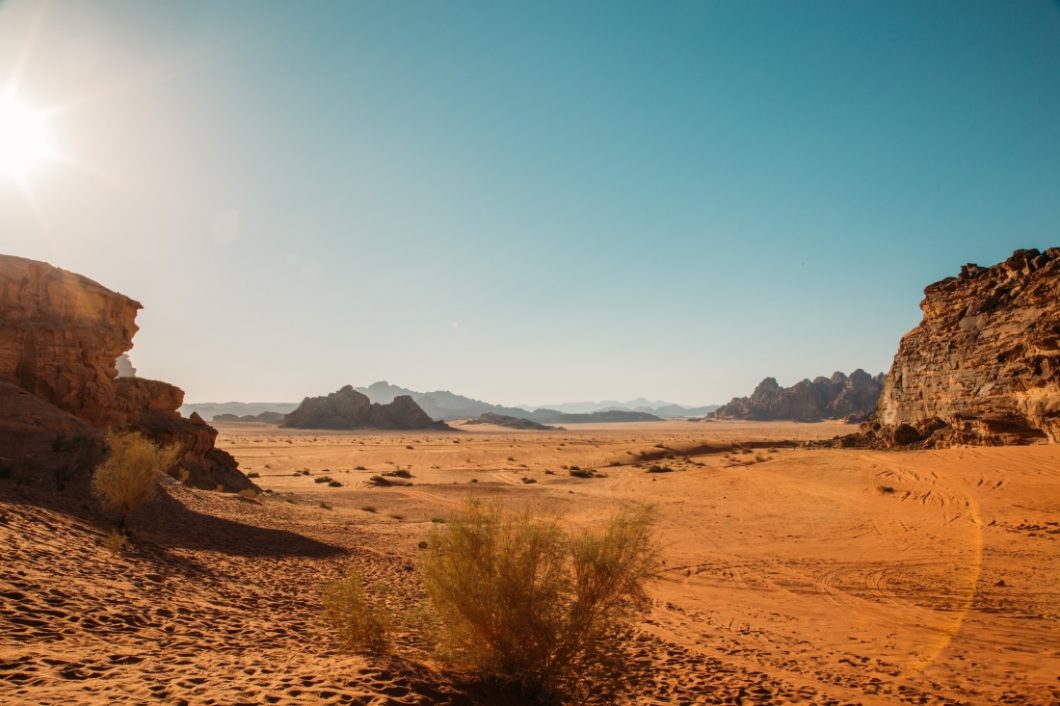 A vast desert landscape in Wadi Rum in Jordan. The sun shines in a clear blue sky overhead.