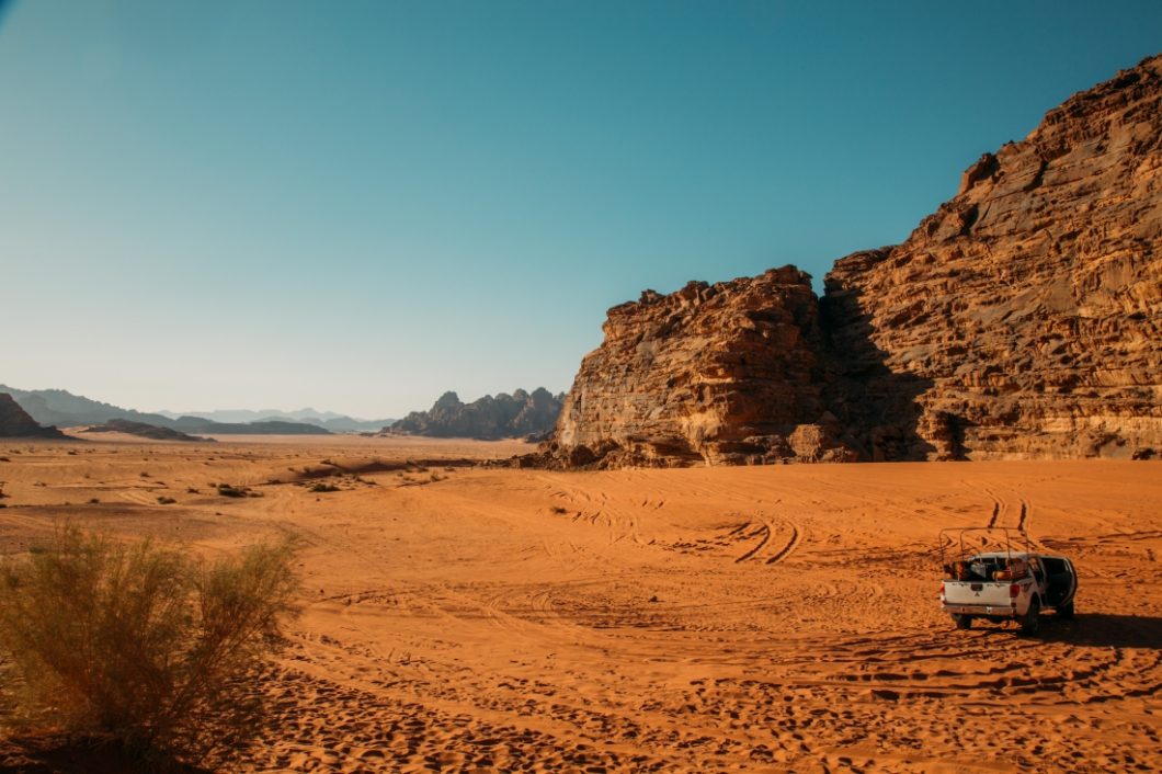 A white four-door pick up truck idles in a Wadi Rum desert valley while tourists admire the rust-red sands and rocky mountains on a clear, sunny day.
