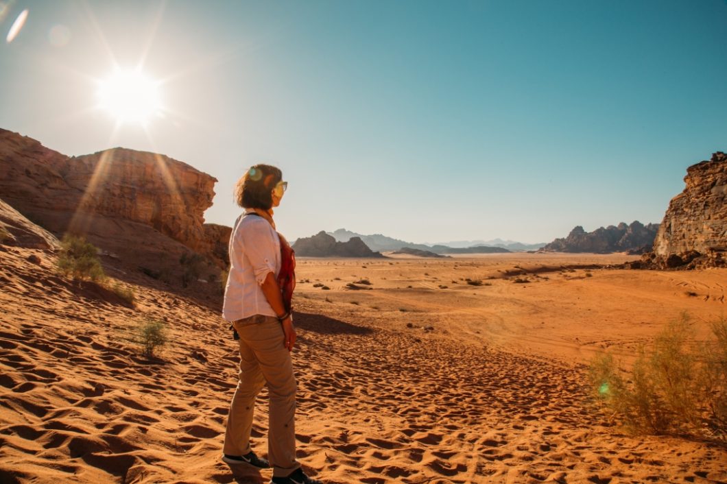 A woman stands on a sandy hill in the Wadi Rum desert on a sunny, clear day. She's wearing a white long-sleeve linen shirt, red scarf, and long khaki hiking pants, and looking out over the desert landscape.