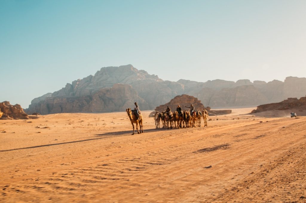 A heard of camels with tourists ride through the sandy valley of the Wadi Rum desert. Rock formations rise against a hazy sky in the background.