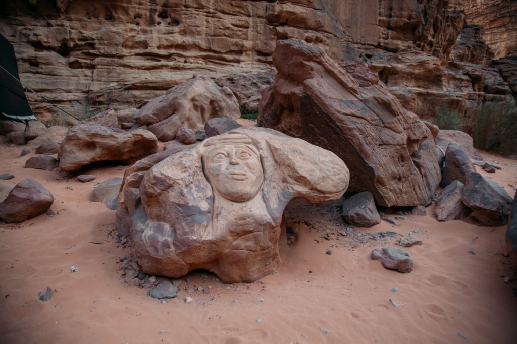 A large boulder in the Wadi Rum desert features an ancient Bedouin carving of a face and bust. The boulder sits among a group of rocks near a mountain in the Wadi Rum desert.