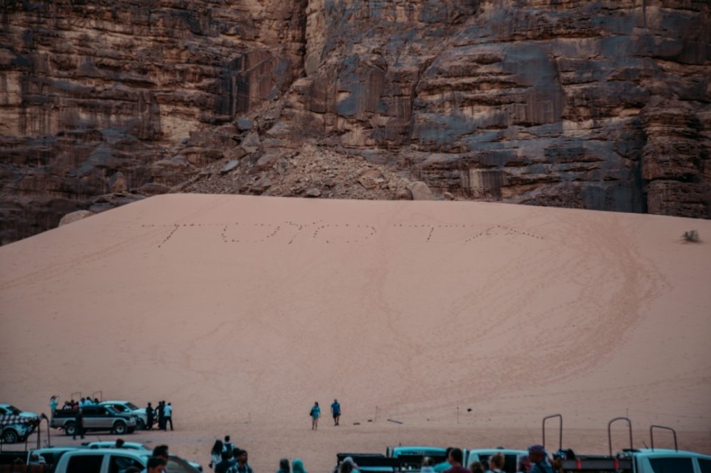 Multiple Jeep tour groups converge at the base of a giant sand dune that rests along a rocky mountain in the Wadi Rum desert. Tire tracks have spelled out the word "TOYOTA" along the side of the sand dune.