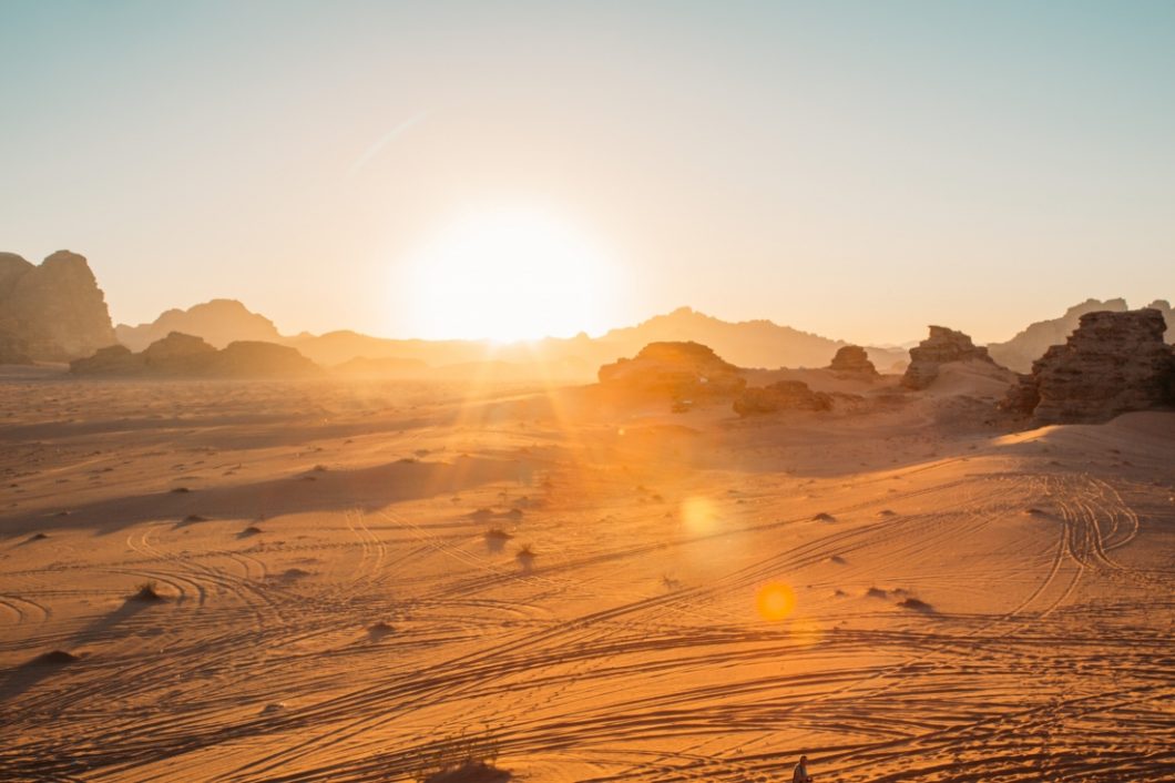The sun raises over the Wadi Rum desert valley, soaking the red sand and rocky mountains in a golden light. Tire tracks from four wheelers and Jeep tours are seen on the valley ground.