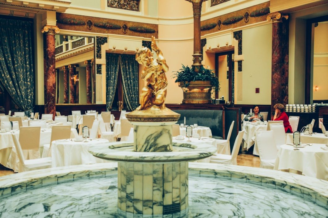 A marbled tiled fountain in the center of a grand ballroom dining room. The fountain features a gold angel statue in the center, and is a key location from the book, A Gentleman in Moscow.