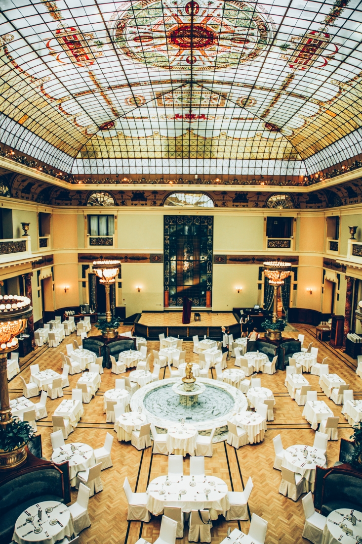 A gorgeous view of the Piazza dining room from a second floor balcony. Dining tables are arranged throughout the room and around a stunning marble fountain. The sun shines in through the vast stained glass ceiling.