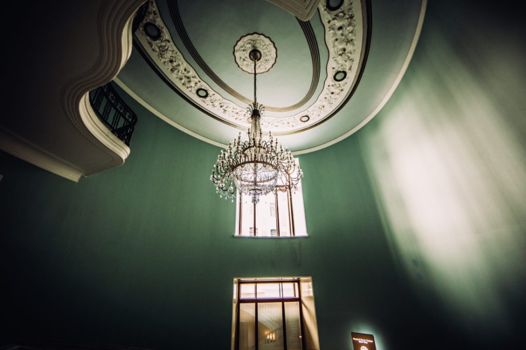 A high ceiling with a crystal chandelier handing down in front of glass windows.