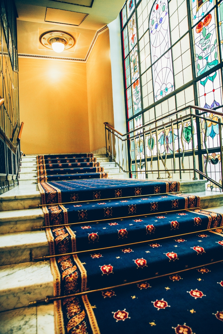 A marble staircase inside The Metropol Hotel. The staircase is lined with an ornate carpet and is naturally lit by a wall of bright stained glass windows.