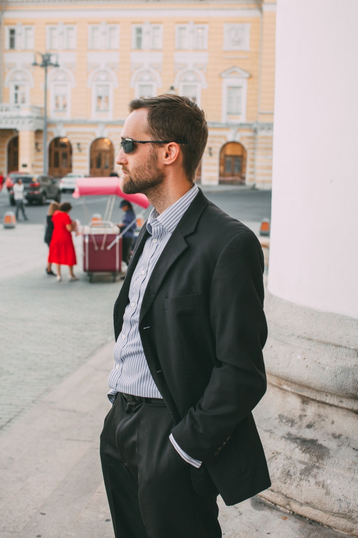 A man stands in a Moscow city street, facing away from the camera for a profile view of his black suit and pinstripe collard shirt.