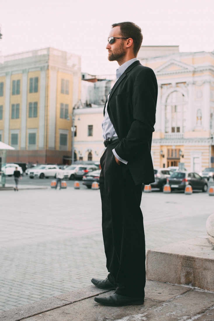 A man poses in a Moscow city square, wearing a black suit jacket and pants paired with a pinstripe button-down shirt and sunglasses.