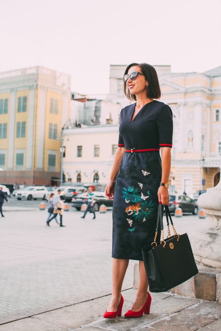 A woman poses standing on the side of a Moscow street outside the Bolshoi theater. She's wearing a black dress with an embroidered calf-length skirt and red pumps. She's holding a large leather purse.