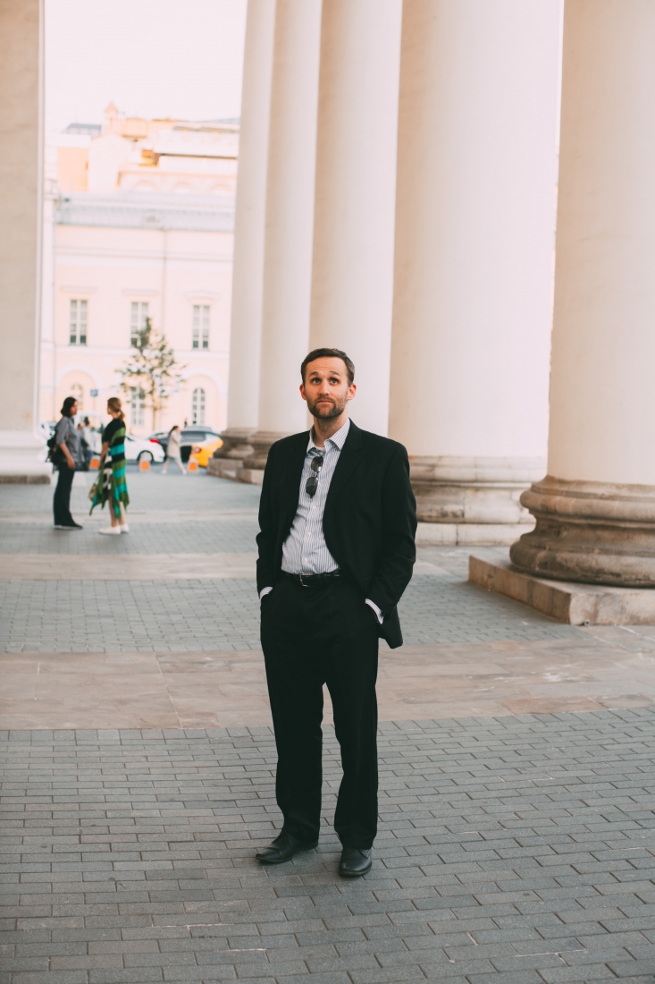 A man stands outside the Bolshoi theater in Moscow, wearing a black suit and blue stripped button-down shirt.