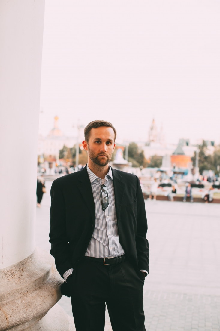 A man stands with his hands in his pockets, looking at the camera and modeling a black suit and pants with a white and blue pinstripe button-down shirt.