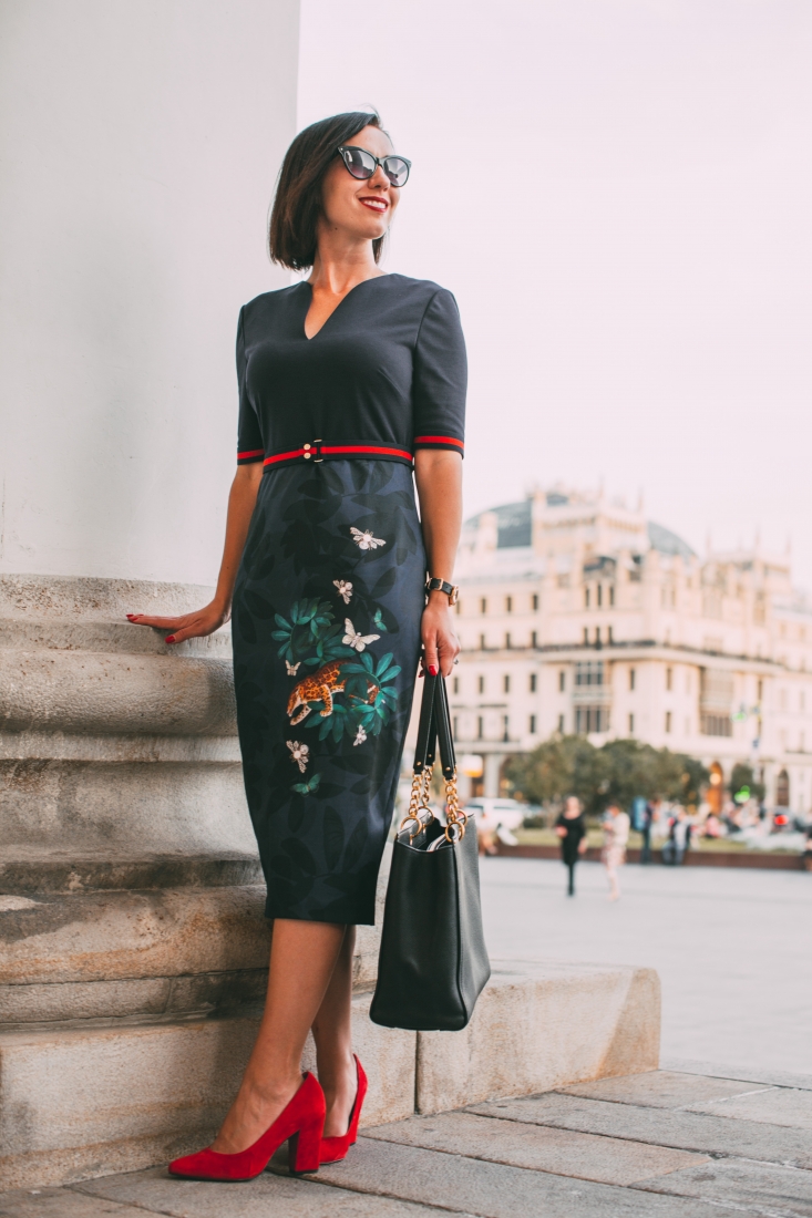 A woman poses next to a stone column in a Russian city square. She's wearing a stylish black dress with an embroidered skirt, red high heeled shoes, and carrying a black leather purse.