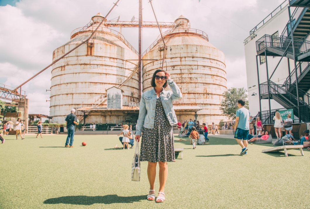 Lindsey wearing a floral grey Karina dress with a denim jacket standing on green turf at Magnolia Market in Texas