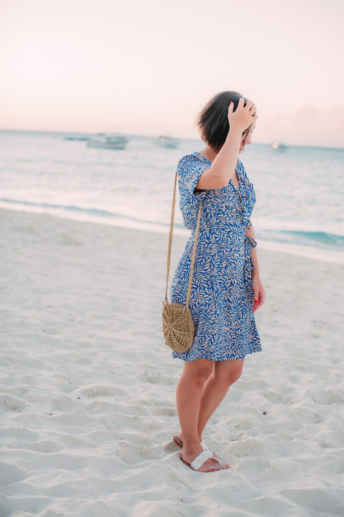 Lindsey wearing a silk wrap dress in a leaf print on the beach, touching her hair