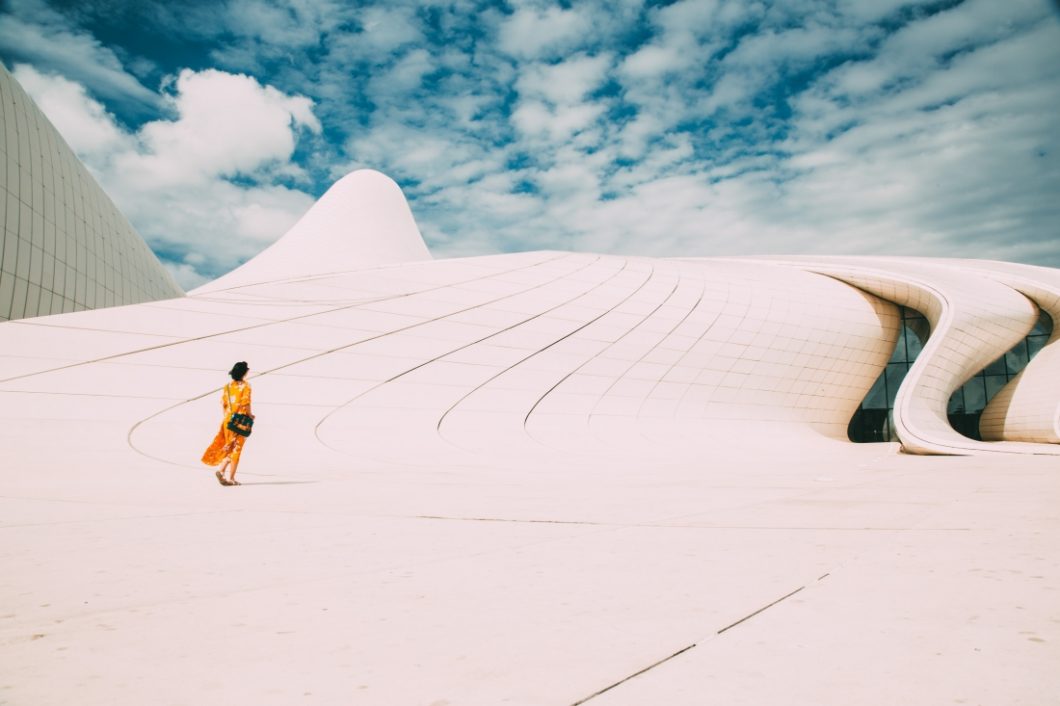 A woman walks along a large concrete walkway alongside a curved concrete building in the city of Baku. The sky is blue and filled with fluffy white clouds. The woman is small in the photo, wearing a yellow dress and carrying an crossbody bag.