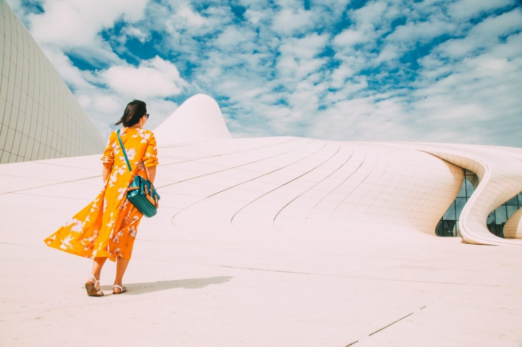 A woman walks along a concrete path next to a curvy concrete building in the city of Baku, Azerbaijan. Her back is turned to the camera and she's wearing a long yellow and white floral maxi dress. The sky is blue and cloudy.