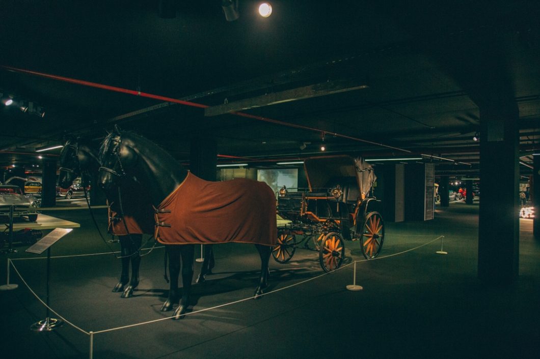 A 1890s Benz Viktoria horse-drawn carriage is displayed with two horses pulling the black carriage body on wheels. 