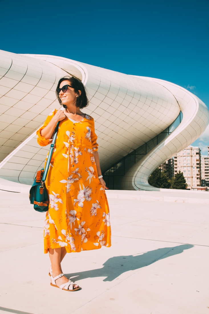 A woman stands on a concrete sidewalk in front of a large, curvy concrete building. She's wearing a long, marigold colored dress with a while flower print, white sandals, and a forest green over-the-shoulder messenger bag.