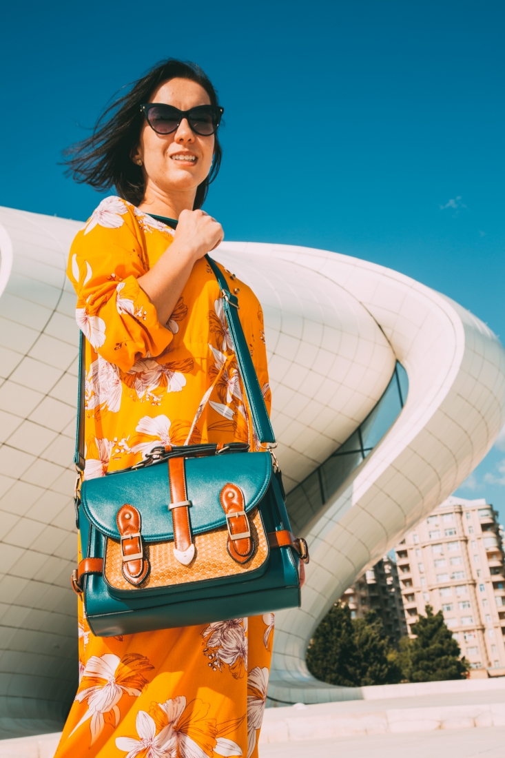 A woman stands in front of a large, curvy concrete building against a clear blue sky in Baku, Azerbaijan. She's wearing a long yellow dress with white floral print, and holding a deep green over-the-shoulder messenger bag with caramel colored buckles.