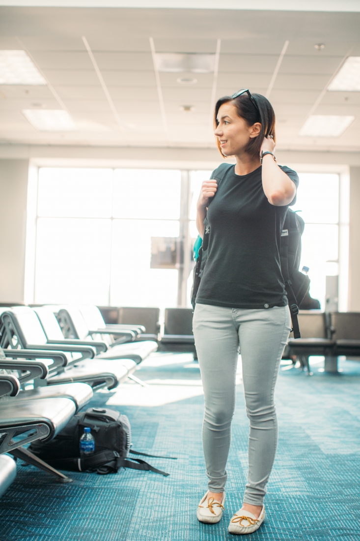 A woman stands in an airport terminal, wearing a charcoal gray t-shirt, light wash jeans, and moccasin shoes, with a travel backpack over her shoulder.