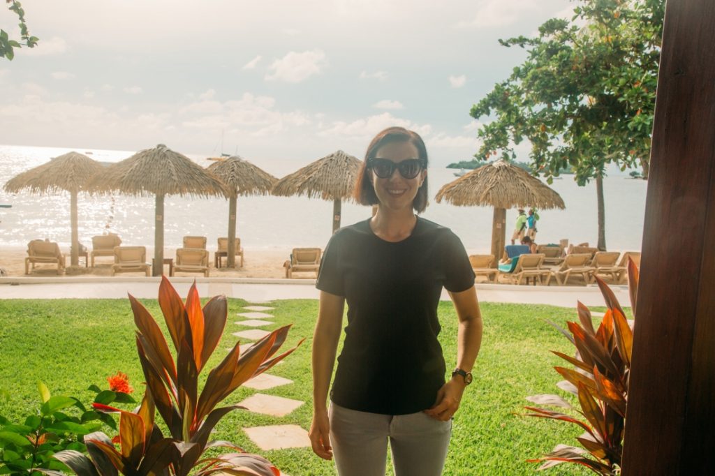 A woman stands in front of a beach in Jamaica, small beachfront huts lined behind her. She's wearing a black t-shirt from the sustainable travel clothing company Aviator.