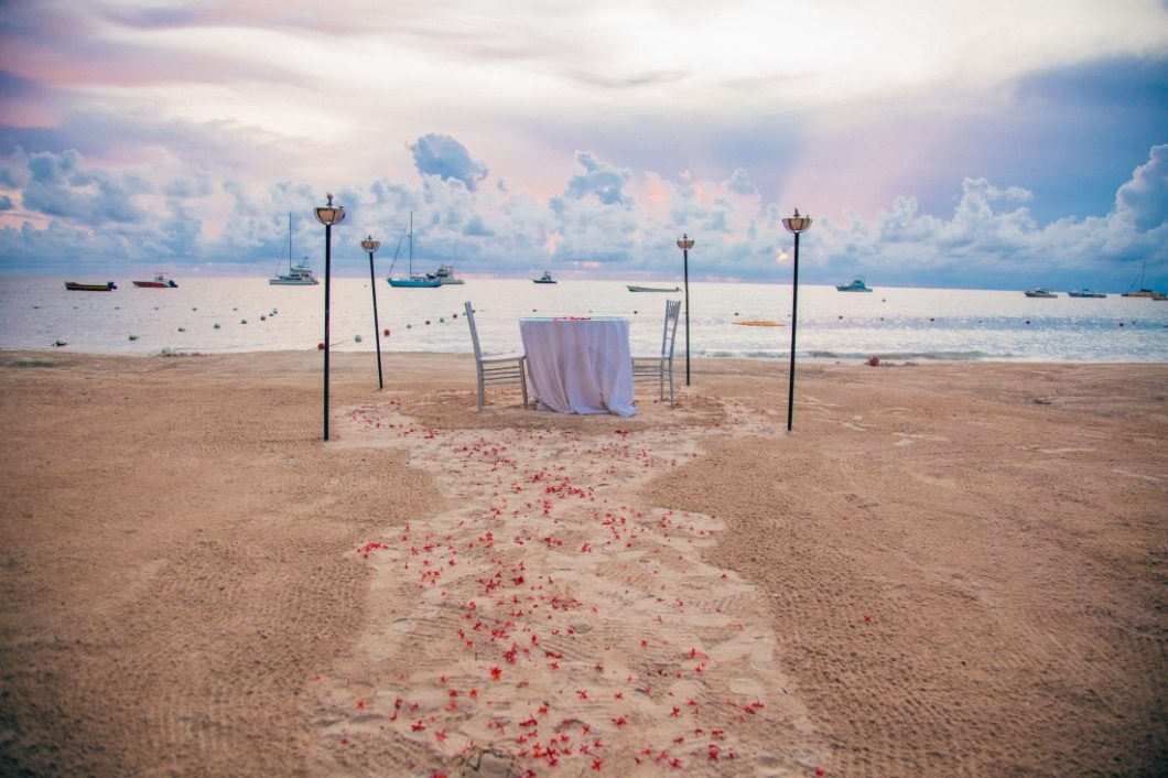 A stunning dinner table for two set up on the beach in Negril, Jamaica. A sweethearts table sits at the end of a sandy path of flower petals, surrounded by lanterns. In the background. boats idle in the ocean that expands into a picturesque cloudy horizon.