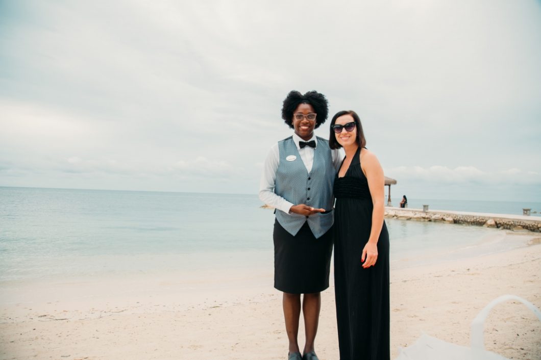 Lindsey wearing a black maxi dress standing next to her butler on the beach who is wearing a Sandals uniform and glasses