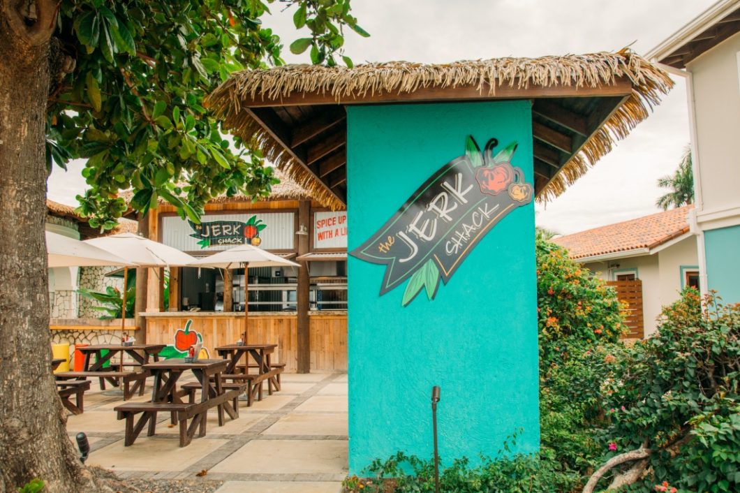 the jerk shack sign on a bright blue wall and sitting area featuring wooden picnic tables and cream umbrellas