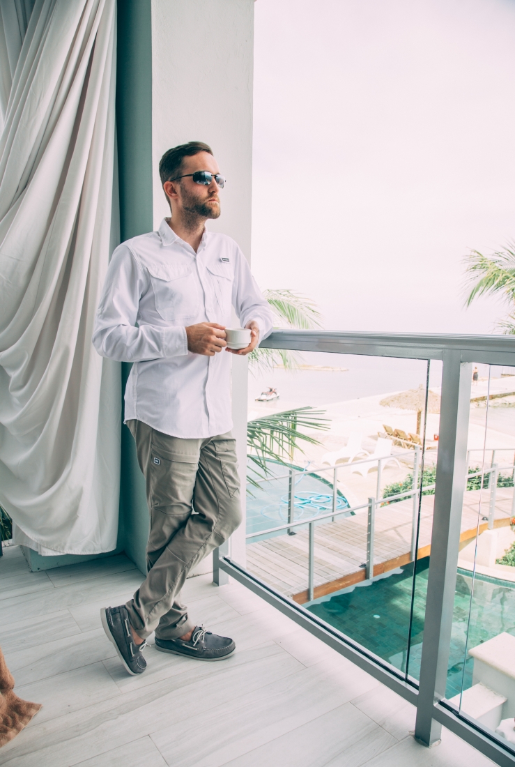Zac wearing sunglasses, a white button down shirt, grey hiking pants, and grey boat shoes, standing on a balcony at the Sandals Montego Bay Resort