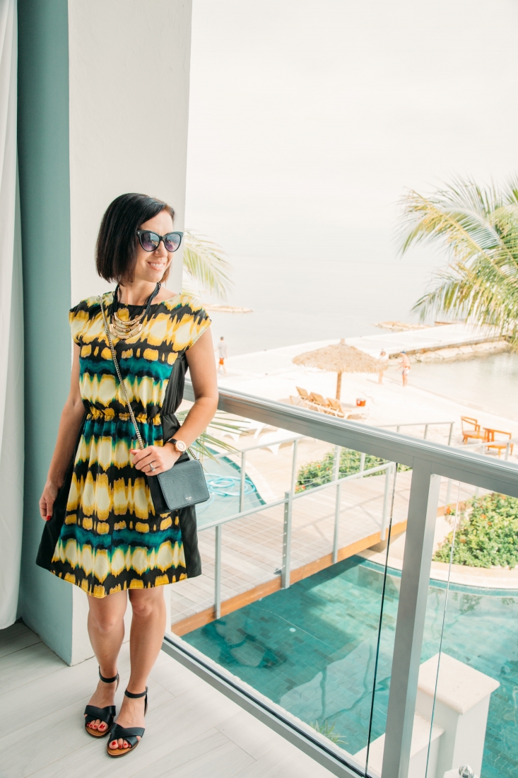 Lindsey is standing on a balcony at Sandals Resorts Montego Bay wearing a black, yellow, and turquoise knee length dress with a large gold and black necklace, black sunglasses and black crossbody purse for her resort evening look