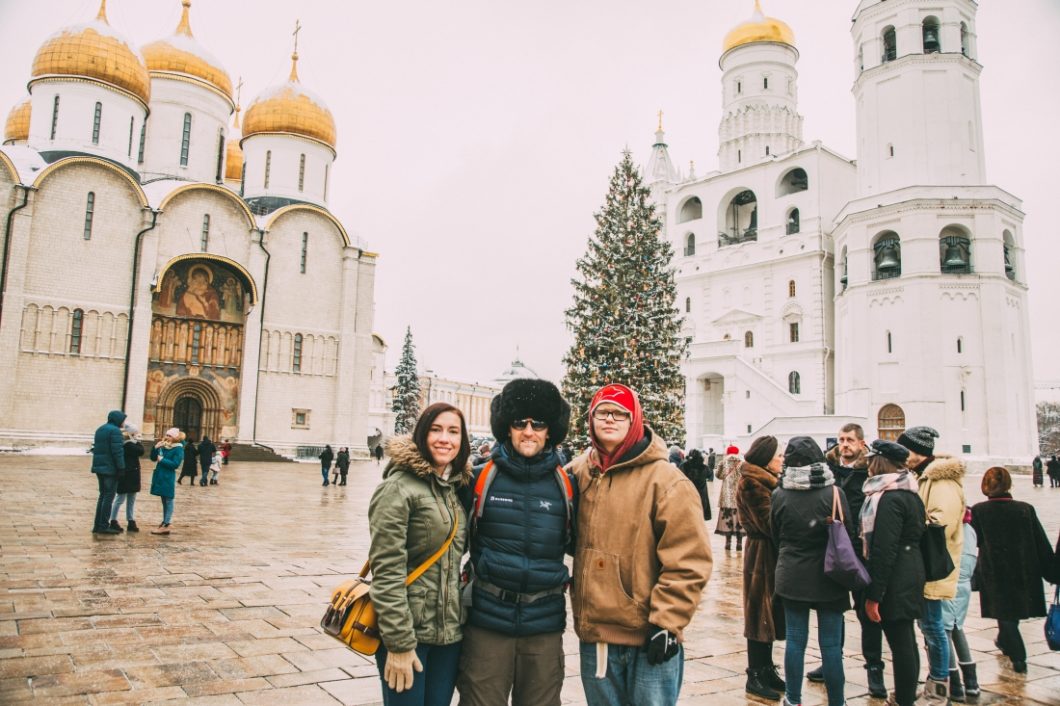 Three people pose in front of large white buildings in a Russian city square. Tourists stand in groups in the background of the photo.