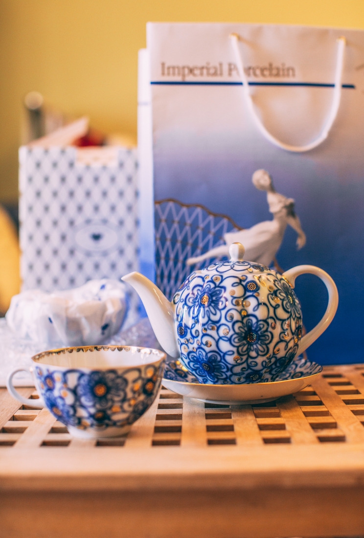 A small tea cup and matching tea kettle sit on a table. The set is made of porcelain and has a blue and gold floral pattern painted across the surface.