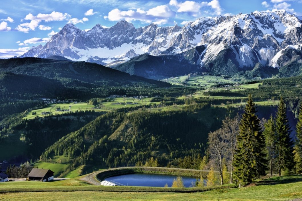 A stunning landscape image of the Italian Alps. A vast green valley filled with hills, trees, and a pond extend back to a snow-covered mountain range and clear blue, cloudy sky.