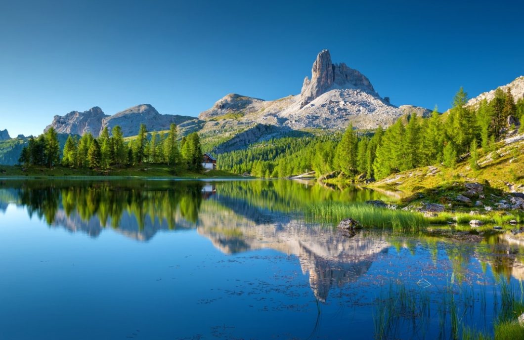 A stunning vista of the Italian Alps. A clear blue lake is lined with a dense forest of trees and hills, which extends to the base of a towering mountain range in the background. The scenery reflects on the calm surface of the lake.